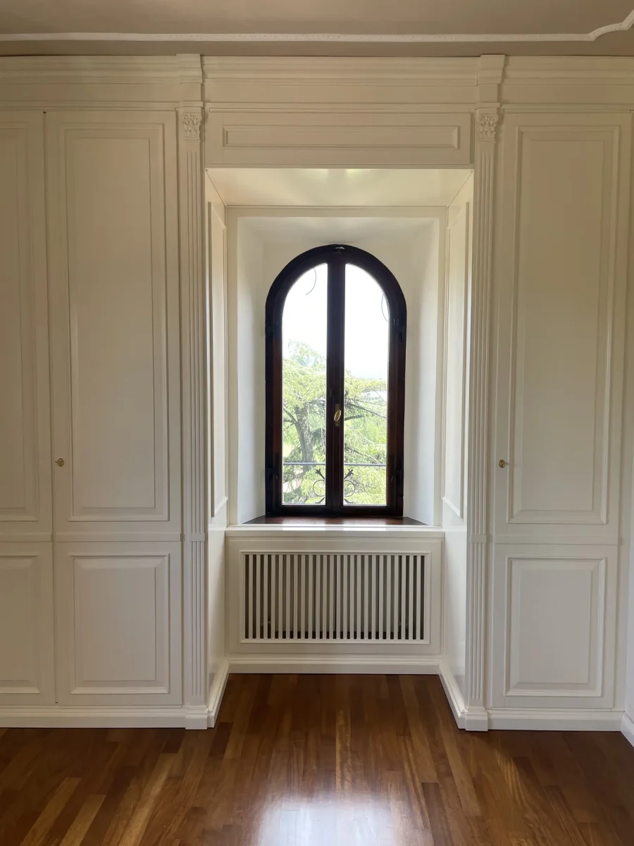 Finestra ad arco con telaio scuro incorniciata dalla boiserie bianca sul parquet