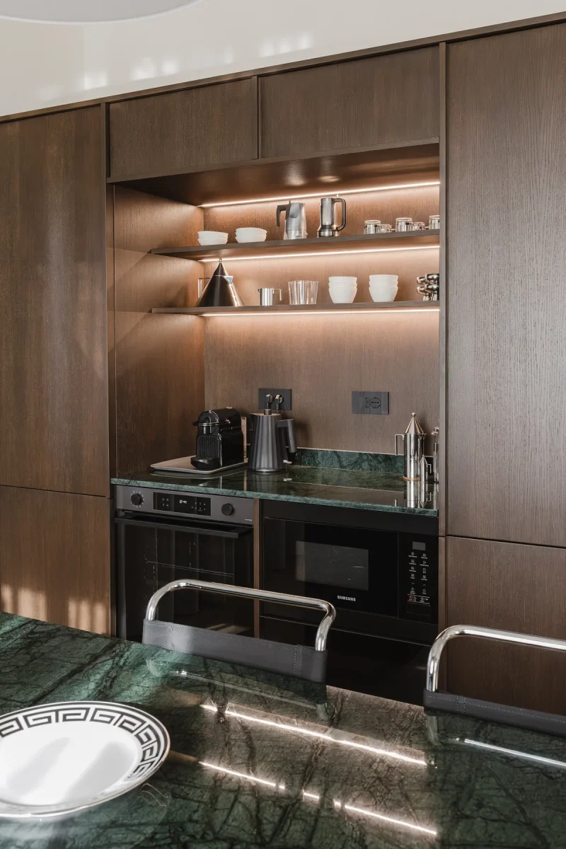 Credenza a giorno in legno wengè con ripiani retroilluminati a LED, stoviglie bianche e caffettiere in acciaio; sotto, forni neri a incasso.