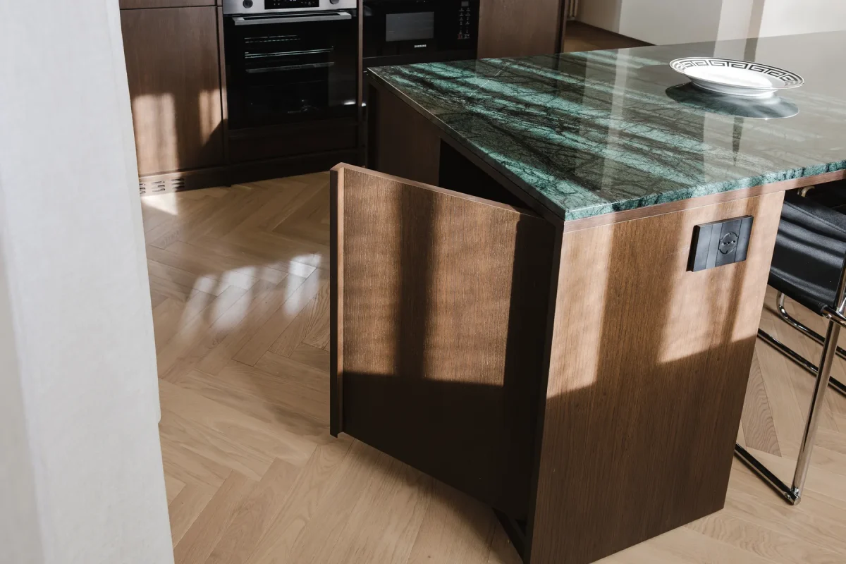Fianco dell'isola in legno wengè con piano in marmo verde alpi, presa a scomparsa e luce radente sul parquet a spina di pesce.
